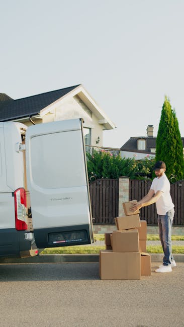 A young man wearing a white t-shirt and dark pants is standing next to an open standard white moving van parked on a residential street. He is holding a cardboard box, preparing to load it into the ve
