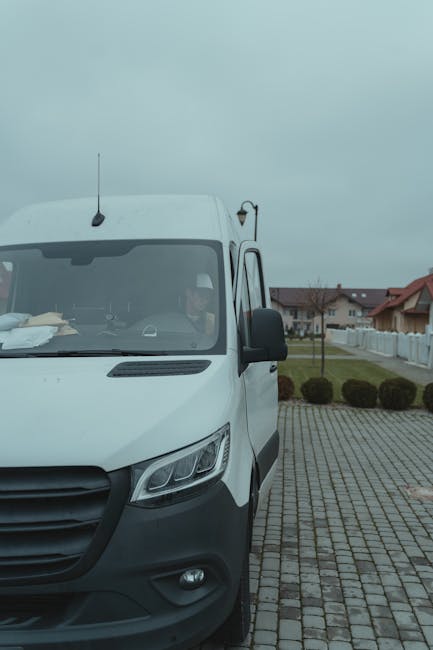 A young man wearing a white t-shirt and dark pants is standing next to an open standard white moving van parked on a residential street. He is holding a cardboard box, preparing to load it into the ve