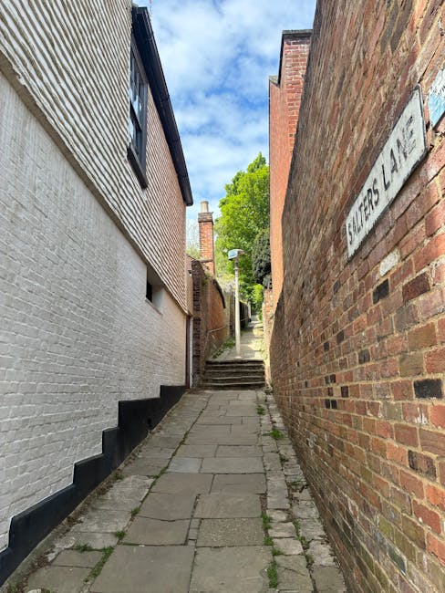 A narrow brick-paved alleyway with a slight upward slope, flanked by a white-painted building on the left and a red-brick wall on the right. The white building features small, rectangular windows, whi