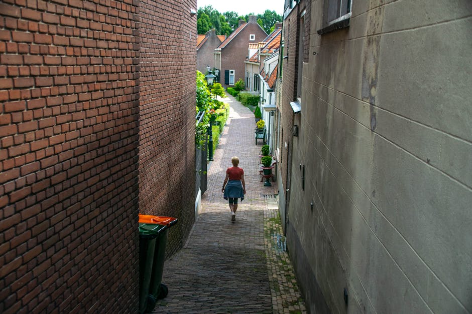 A narrow brick-paved alleyway with a slight upward slope, flanked by a white-painted building on the left and a red-brick wall on the right. The white building features small, rectangular windows, whi