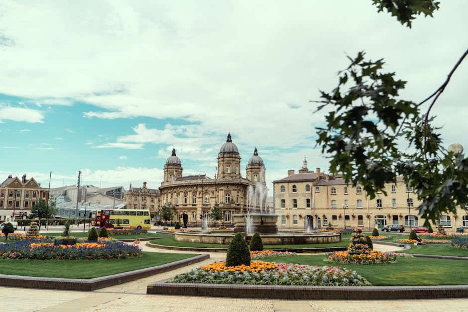 A panoramic view of a historic city square featuring a large ornate stone building with domed towers in the background under partly cloudy skies. In the foreground, there is a circular fountain with multiple water jets spraying into the air, surrounded by neatly landscaped flowerbeds with colorful blooms and trimmed bushes. To the left, a yellow double-decker bus is seen parked on the street near other vehicles, while a paved pavement runs through the square. In the upper right corner of the image, a branch of a leafy green tree partially extends into the frame. This scene depicts a vibrant, well-maintained urban area suitable for home relocation and furniture transport, with elements typical of a town centre move and logistics managed by professional removals services like Removal Company Kingston.