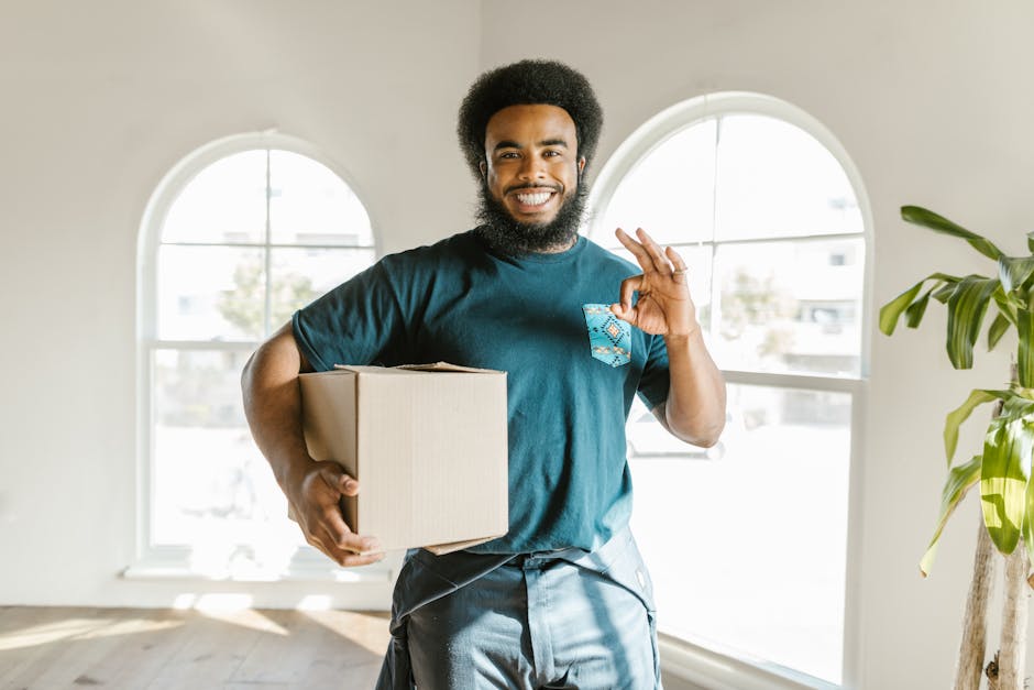 A smiling man with a dark beard and curly hair stands behind several packed cardboard moving boxes inside a well-lit room with a large arched window. The boxes, some sealed with red tape and others wi